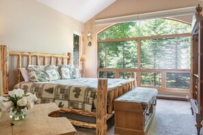 A bedroom with a large window, a wooden bed, a bench, and a small table with a vase of flowers.