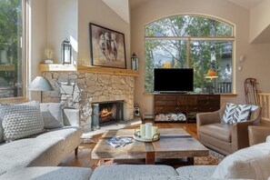 Una sala de estar acogedora con chimenea de piedra, asientos cómodos, un televisor de pantalla plana y un ventanal con vistas a árboles.