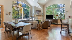Amplia sala de estar con techo alto, ventanas grandes que ofrecen vistas a árboles, comedor con mesa y sillas de madera, y zona de estar acogedora con sofás y chimenea.