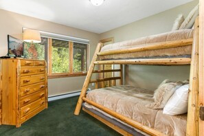Uma cama beliche com escada de madeira, um penteou com várias gavetas e uma janela com vista para o verde.