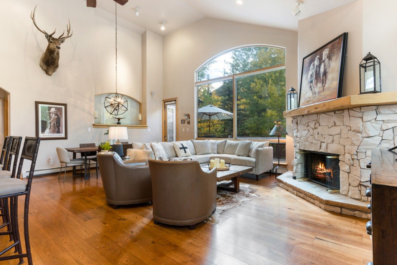 A spacious living room with a stone fireplace, a mounted deer head, and a large window offering a view of trees.