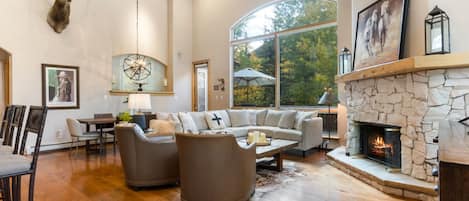 A spacious living room with a stone fireplace, a mounted deer head, and a large window offering a view of trees.