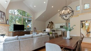 Un amplio salón con un sofá seccional grande, una mesa de comedor con sillas, una araña y una zona de cocina visible a través de un arco abierto.