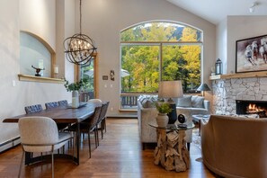 Uma sala de estar espaçosa com lareira, área de jantar com uma mesa grande e vista para árvores através de uma janela ampla.