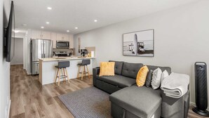 Un espacio habitable moderno con un sofá gris, una pequeña zona de cocina y un televisor de pantalla plana montado en la pared.
