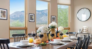 A dining table set with wine glasses, a loaf of bread, and a vase of flowers, with mountain views through large windows.