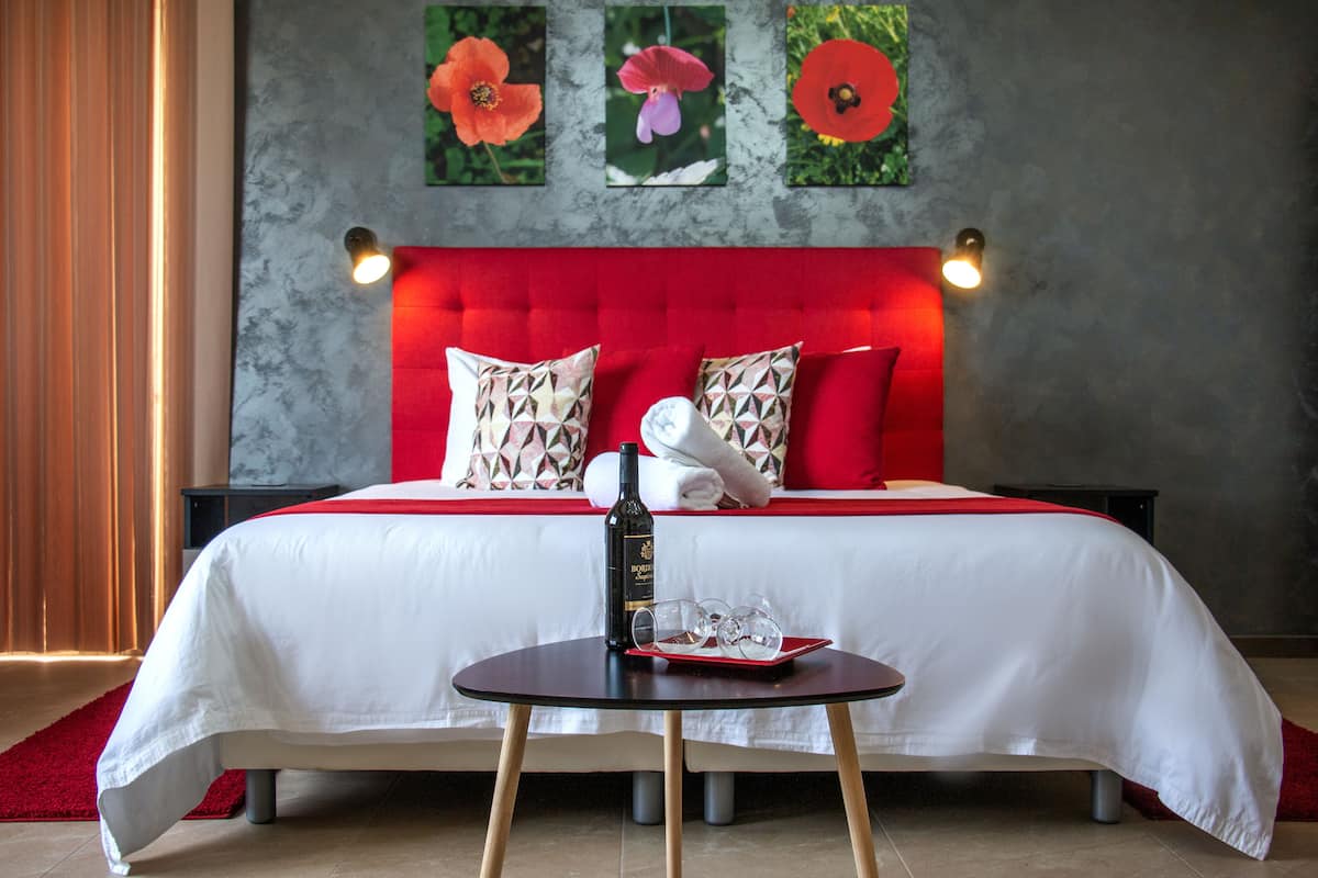 Ein Hotelzimmer mit einem roten, gepolsterten Kopfteil, einer weißen Bettdecke und einem kleinen Tisch mit einer Flasche und Gläsern.