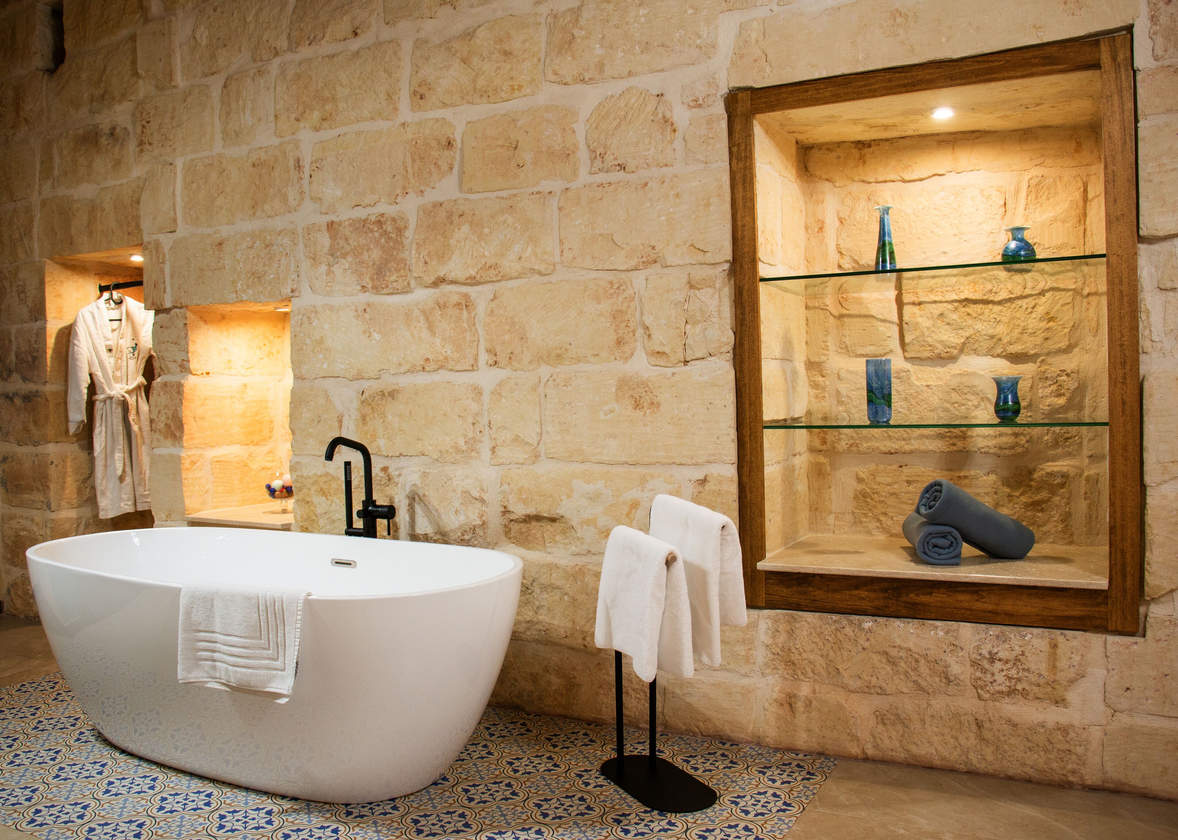 Une salle de bain avec une baignoire sur pieds, une étagère en verre ornée d’objets décoratifs et un mur en pierre.
