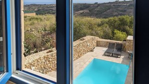 A view from a window overlooking a pool and a landscape with rocky terrain and sparse vegetation. - Sea Holly Gozo (Nadur)