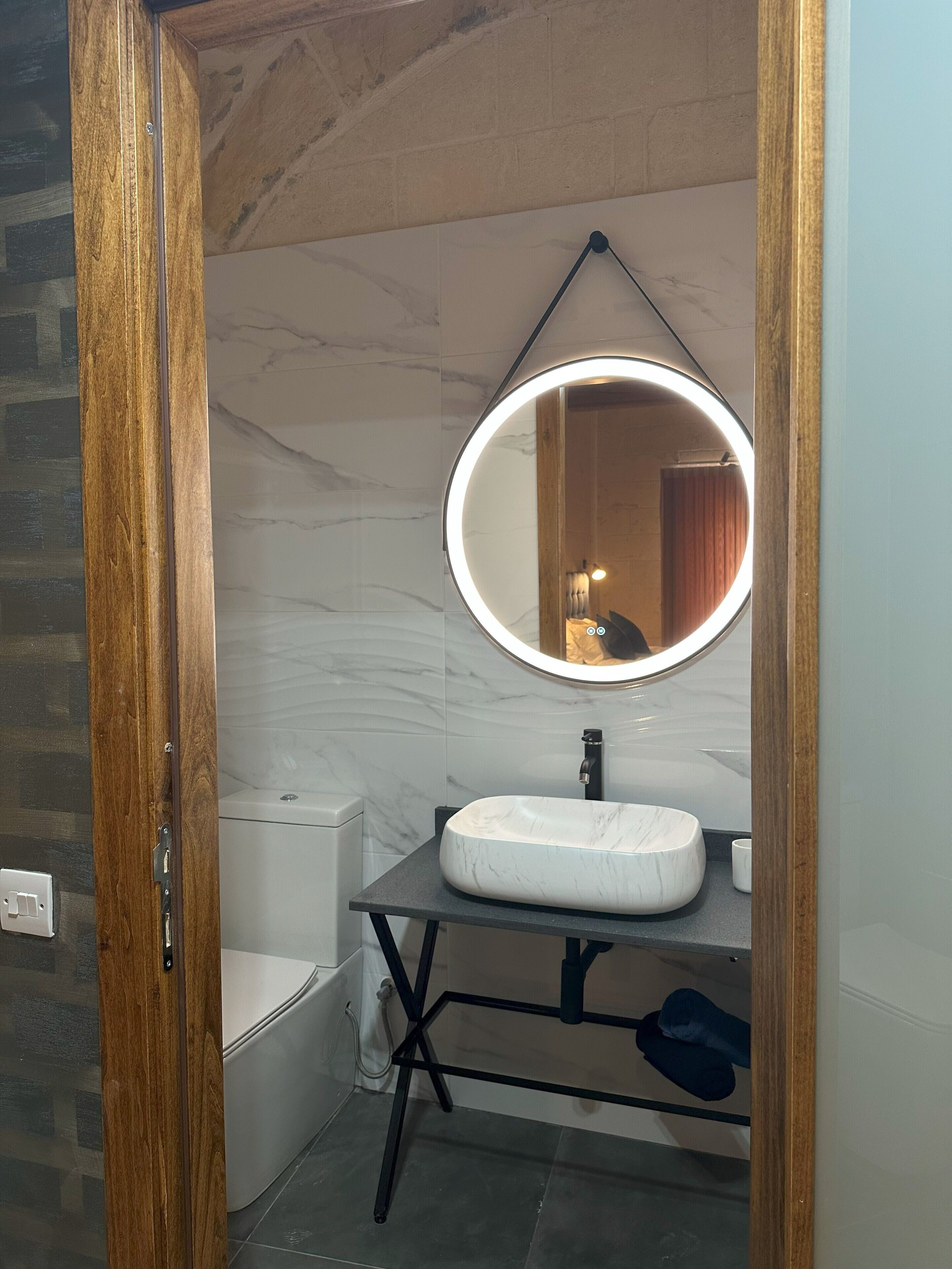 Un baño con pared de mármol, un lavamanos blanco, un espejo con luz circular y un marco de puerta de madera.