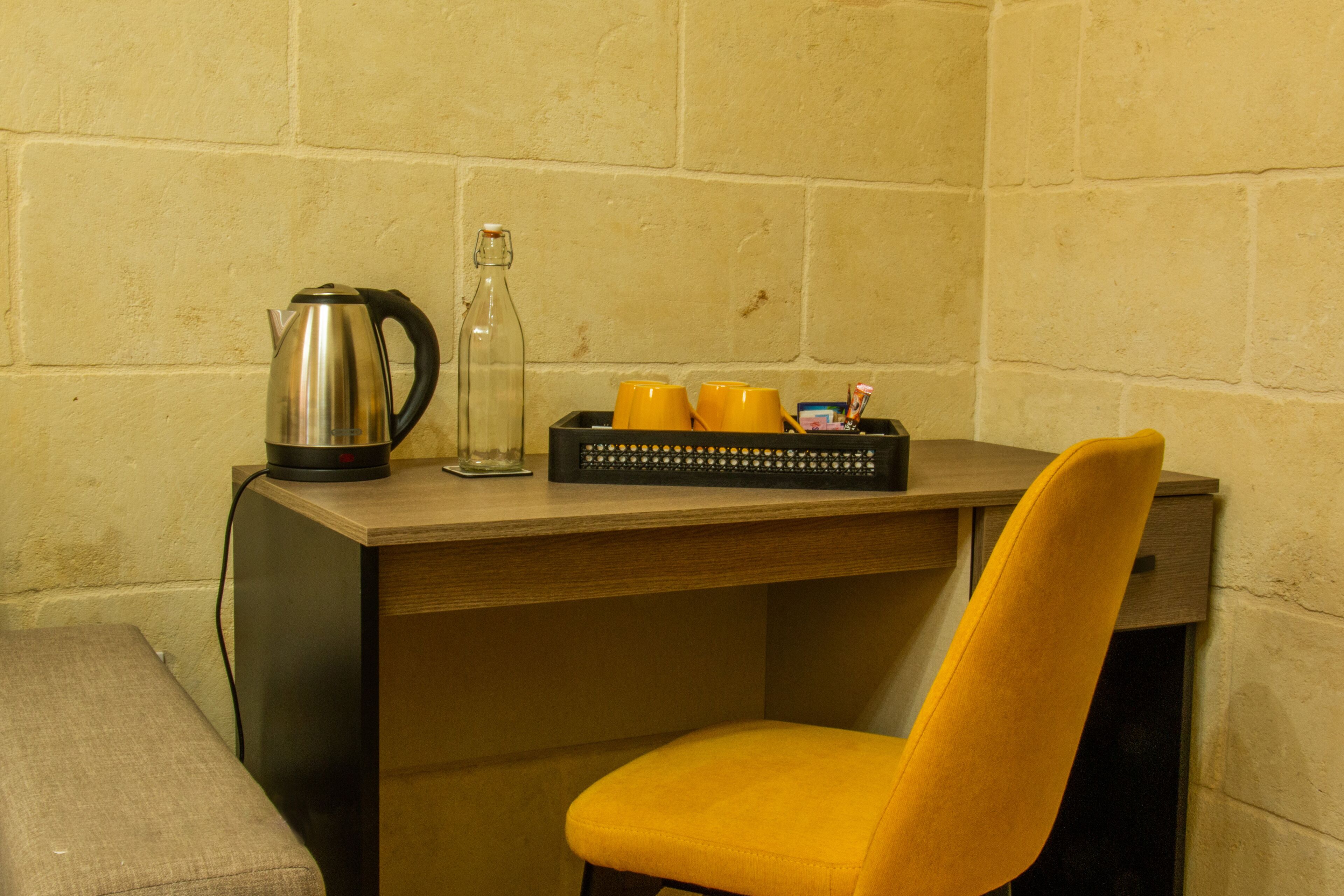 A small, compact room with a wooden desk, a yellow chair, a kettle, a glass bottle, and a tray with cups and a small box.