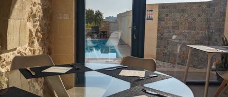 Un área de comedor al aire libre con una mesa y sillas de vidrio, una piscina al fondo y una pared de piedra.