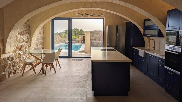 A modern kitchen with an arched stone ceiling, a central island, and a view of a swimming pool. - Sea Holly Gozo (Nadur)