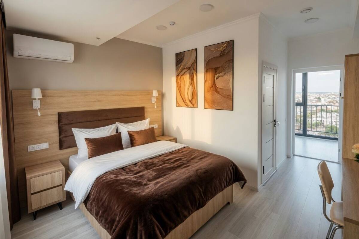 A modern bedroom with a wooden headboard, a large bed, bedside tables, a desk, and a view through a large window.
