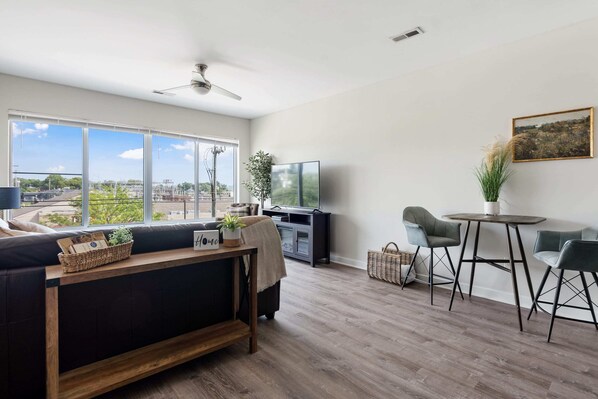 A modern living room with a large window, a ceiling fan, a TV, and a dining area with chairs and a table.