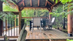 A wooden deck with a thatched roof structure, surrounded by lush greenery.