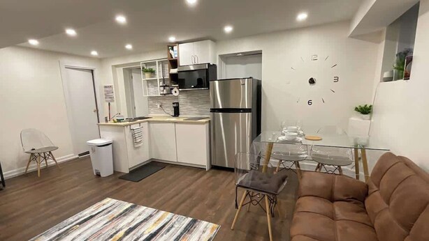 A modern kitchen with a stainless steel refrigerator, a microwave, and a sink. There is a dining table with chairs and a sofa in the living area. - Modern Basement Studio w/ Kitchen, Free Parking (Montréal)