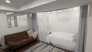 A bedroom with a brown sofa, a bed with a white bedspread, a large window with sheer curtains, and a wooden floor. - Modern Basement Studio w/ Kitchen, Free Parking (Montréal)