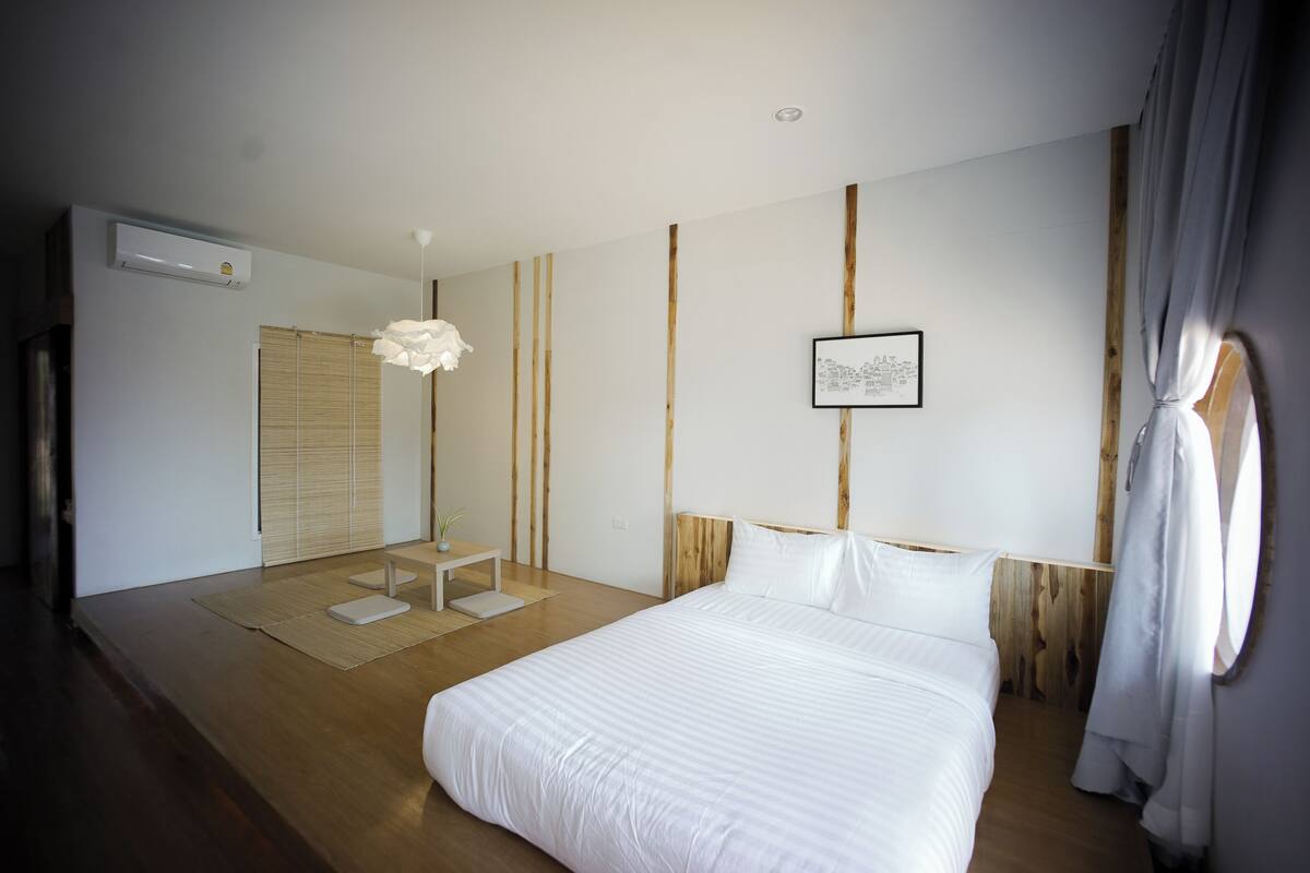A bedroom with a bed, a bedside table, a hanging light fixture, a window with curtains, and a wall-mounted air conditioner.