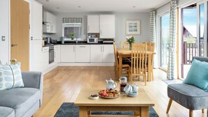 A modern kitchen with white cabinets, a dining area with a wooden table and chairs, and a cozy seating area with a grey sofa and a blue pillow. - Sunbow at Retallack Resort (St. Columb Major)
