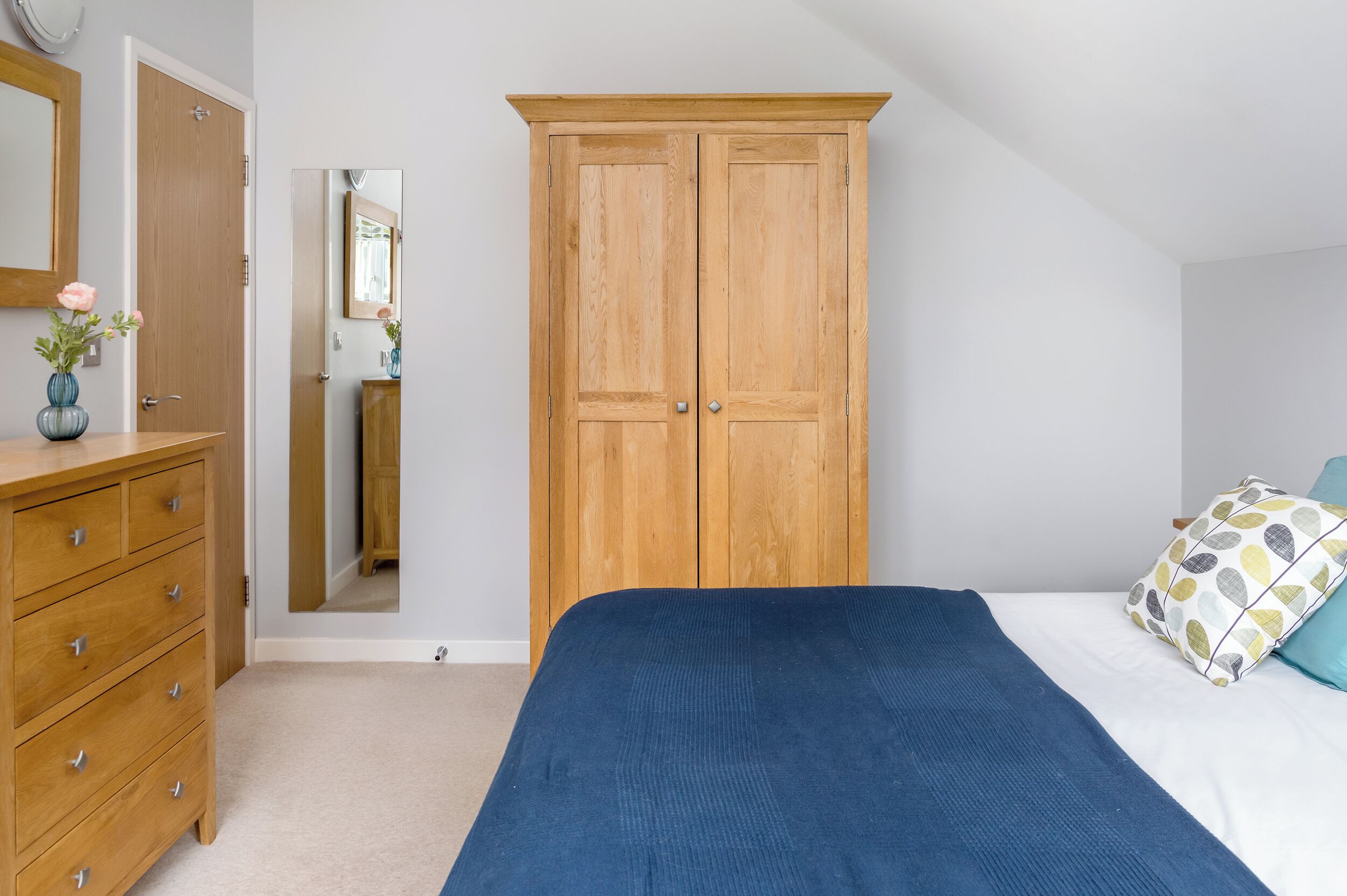 Una camera da letto con un armadio in legno, un letto con coperta blu, una cassettiera di legno e uno specchio.