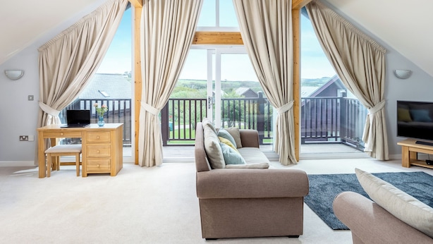 A spacious loft bedroom with a large window, a wooden desk, a sofa, and a TV. - Sunbow at Retallack Resort (St. Columb Major)