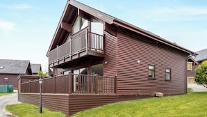 A two-story wooden cabin with a balcony on the upper floor, a fenced front yard, and a clear sky. - Sunbow at Retallack Resort (St. Columb Major)