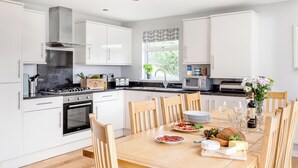 A modern kitchen with white cabinets, a wooden dining table set with plates, glasses, and a bottle of wine. - Sunbow at Retallack Resort (St. Columb Major)