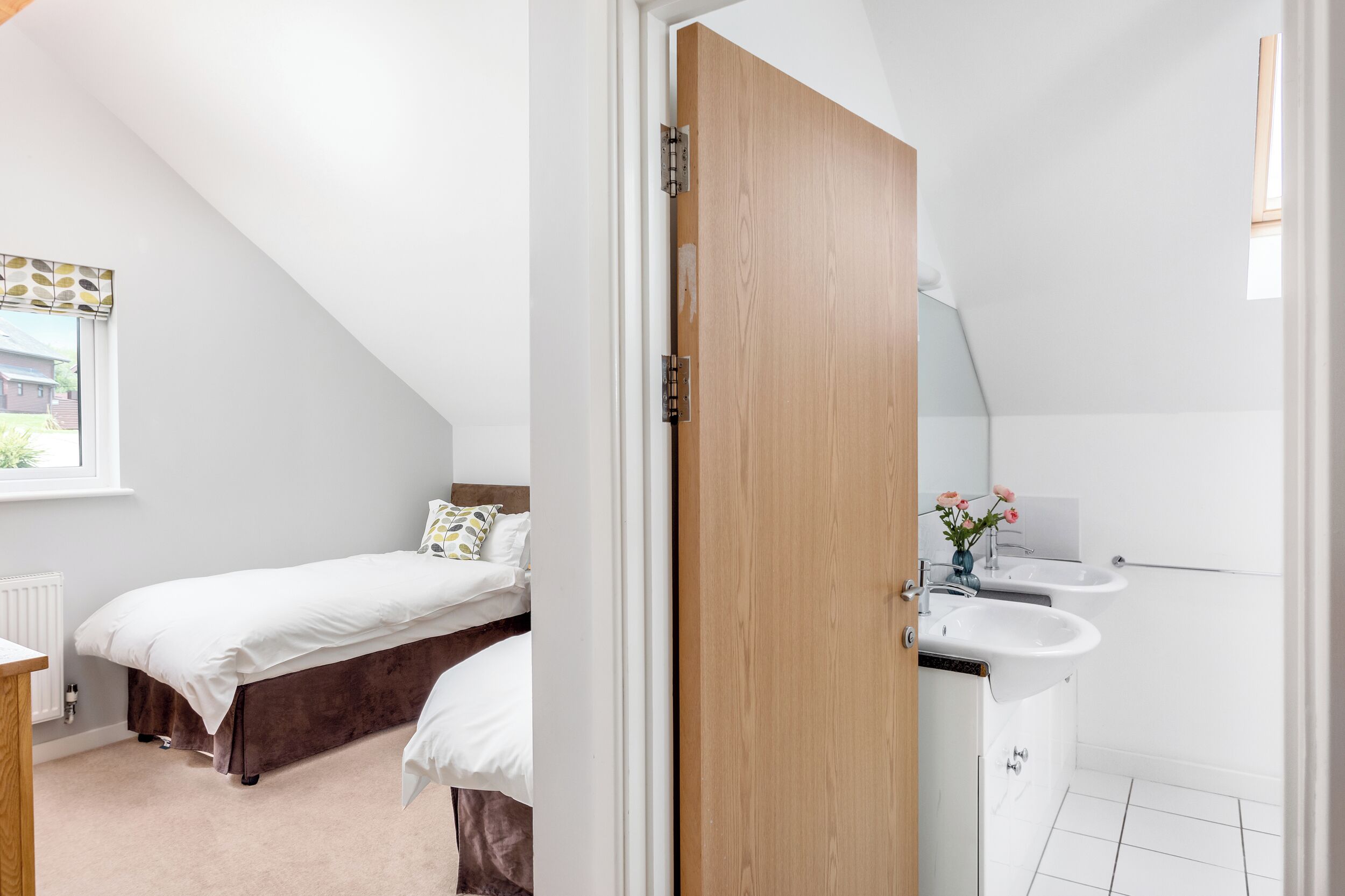 A bedroom with a single bed, a wooden door, a window with a view, and a bathroom with a sink and mirror.