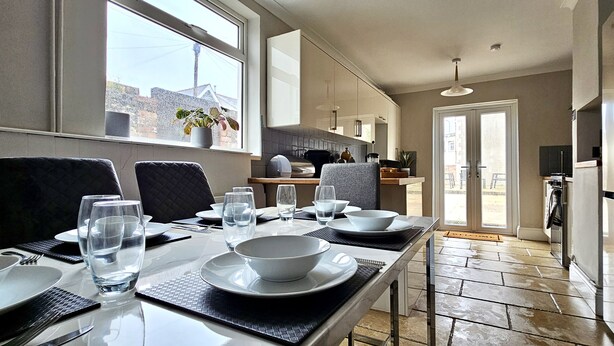 Un coin repas avec une table dressée pour quatre personnes, une cuisine en arrière-plan et une vue sur l’extérieur par une fenêtre.