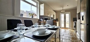 Un coin repas avec une table dressée pour quatre personnes, une cuisine en arrière-plan et une vue sur l’extérieur par une fenêtre.