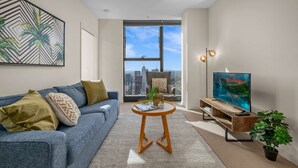 Un salón moderno con vistas a la ciudad, un sofá azul, una mesa de centro redonda de madera y un televisor sobre un mueble de madera.