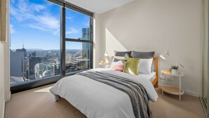 Un dormitorio moderno con un amplio ventanal que ofrece vistas a la ciudad, una cama con muchos almohadones y una mesita de noche con una lámpara.