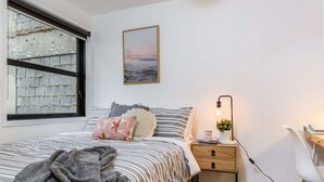 Ein Schlafzimmer mit Bett, Nachttisch, Lampe und Blick auf eine Ziegelwand durch ein Fenster.