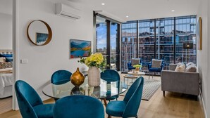 A modern living room with a glass dining table, blue velvet chairs, a grey sofa, and a large window offering a city view. - Premium Chic: A Relaxing Retreat (Port Melbourne)