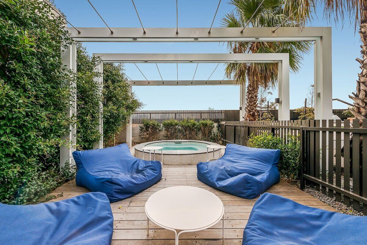 A wooden deck with blue cushions and a small table, leading to a circular hot tub under a pergola.