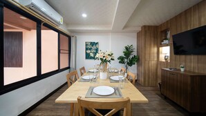 Un coin repas avec une table en bois dressée pour quatre personnes, entourée de chaises. On y trouve une grande fenêtre avec des stores, un tableau accroché au mur et un téléviseur fixé au mur.