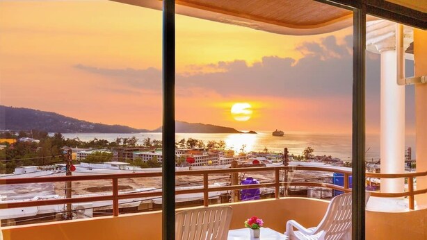 Un balcon offrant une vue sur le coucher de soleil sur la mer, des chaises en plastique blanches et une petite table agrémentée d'une fleur.