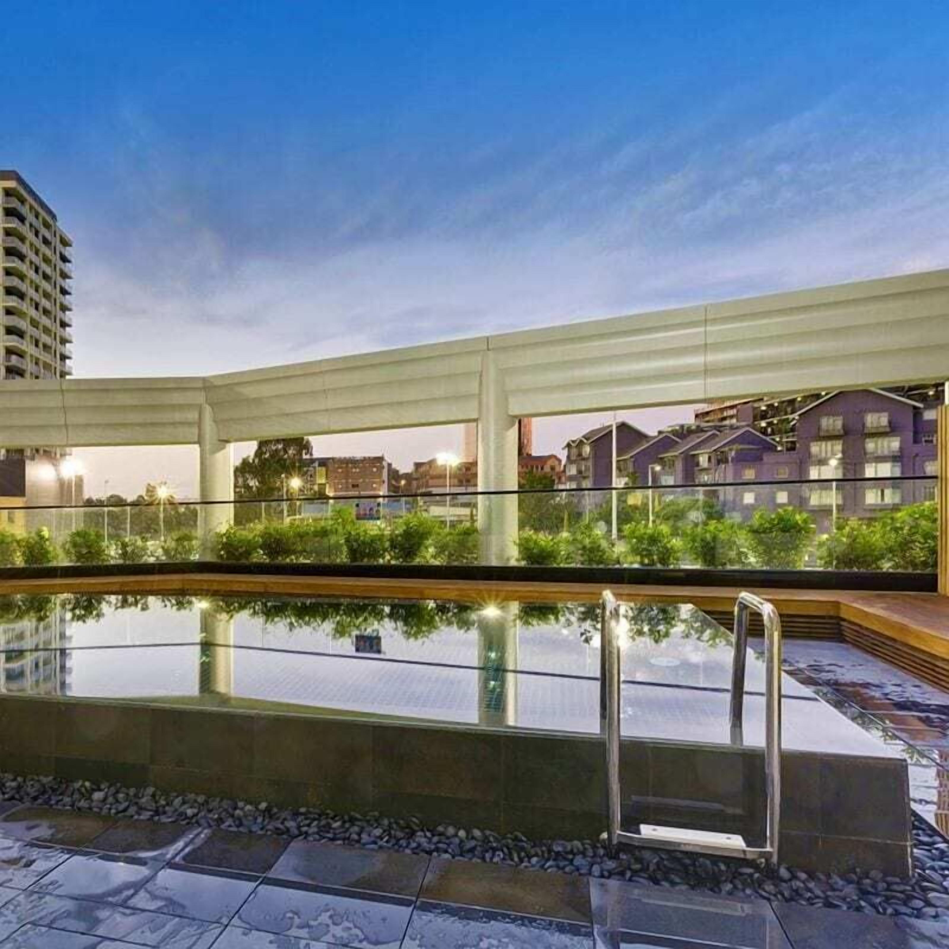 An outdoor swimming pool with a glass barrier, surrounded by greenery and a view of buildings in the background.