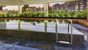 Una piscina al aire libre con barandilla de cristal, rodeada de vegetación y vistas a edificios al fondo.