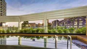 An outdoor swimming pool with a glass barrier, surrounded by greenery and a view of buildings in the background.