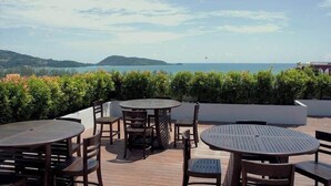 An outdoor patio with round wooden tables and chairs, surrounded by greenery, overlooking a body of water and distant hills. - ✈Sea-view 80sqm-1 Bedroom Home-Patong Beach (Tambon Patong)