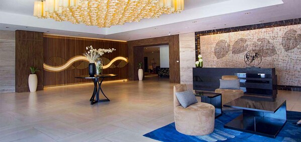Un vestíbulo moderno con una gran lámpara de candelabro, una recepción, un área de estar y una pared con diseño decorativo.