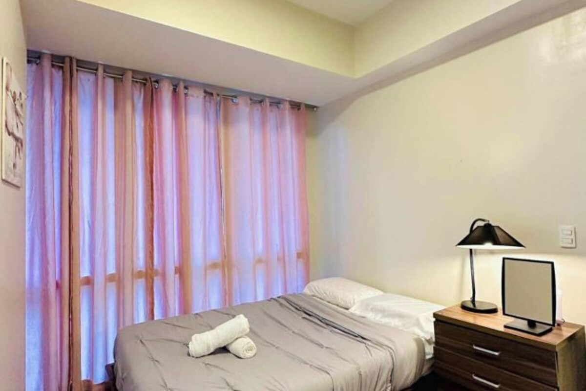 Un dormitorio con cama, mesita de noche con lámpara y cortina.