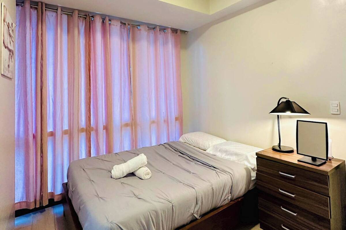 Un dormitorio con cama, mesita de noche con lámpara, espejo y ventana con cortinas.
