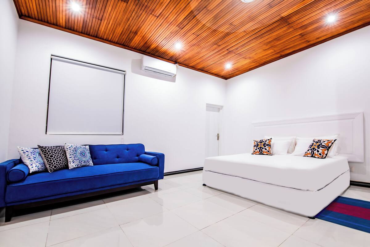 A modern bedroom with a blue sofa, white bed, wooden ceiling, and white walls.