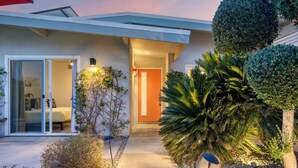 A modern house with a glass door entrance, a concrete pathway, and a well-maintained garden with various plants. - Retro Oasis w/ Pool, Ping Pong, & Large Yard | Lloyd by AvantStay Permit#3779 (Palm Springs)