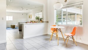 A modern kitchen with a central island, a dining area with orange chairs, and a large window overlooking a garage. - Retro Oasis w/ Pool, Ping Pong, & Large Yard | Lloyd by AvantStay Permit#3779 (Palm Springs)