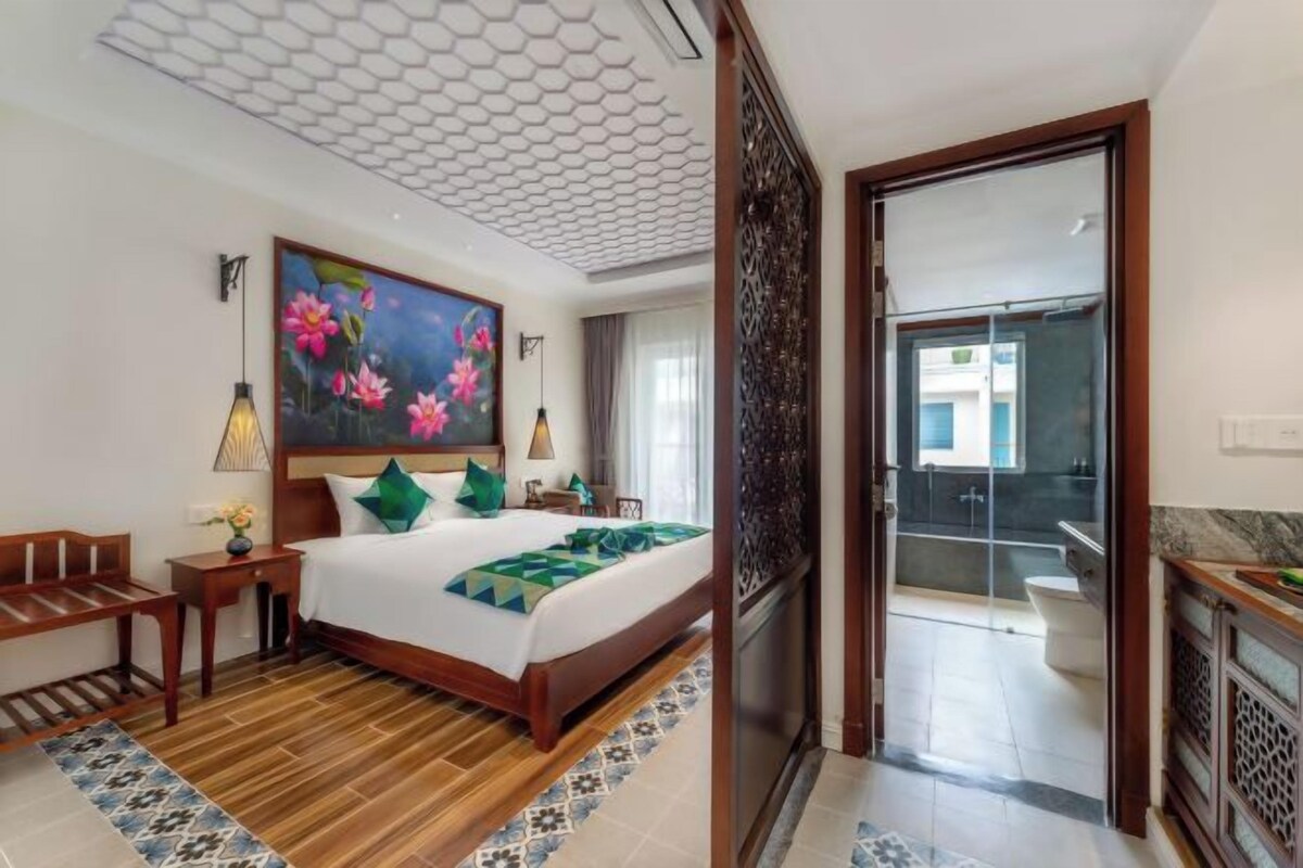 Une chambre d’hôtel avec un lit en bois, un tableau représentant des fleurs, un bureau et une salle de bain visible à travers une porte ouverte.
