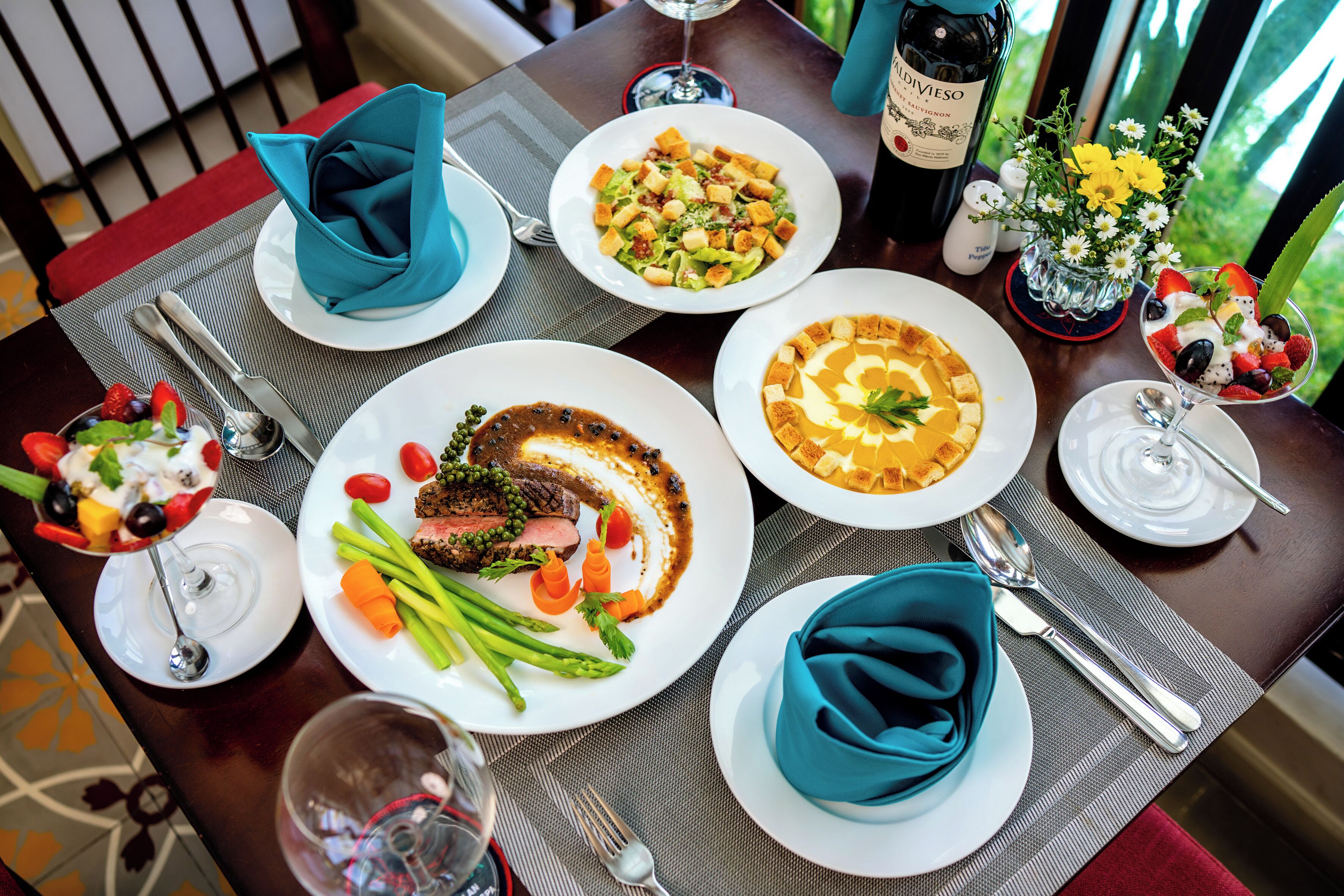 A table set with various dishes, including a salad, steak, and dessert, along with wine and a vase of flowers.